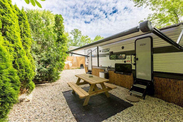 Un camper con tavolo da picnic e zona barbecue, immerso nella vegetazione e circondato da ghiaia.