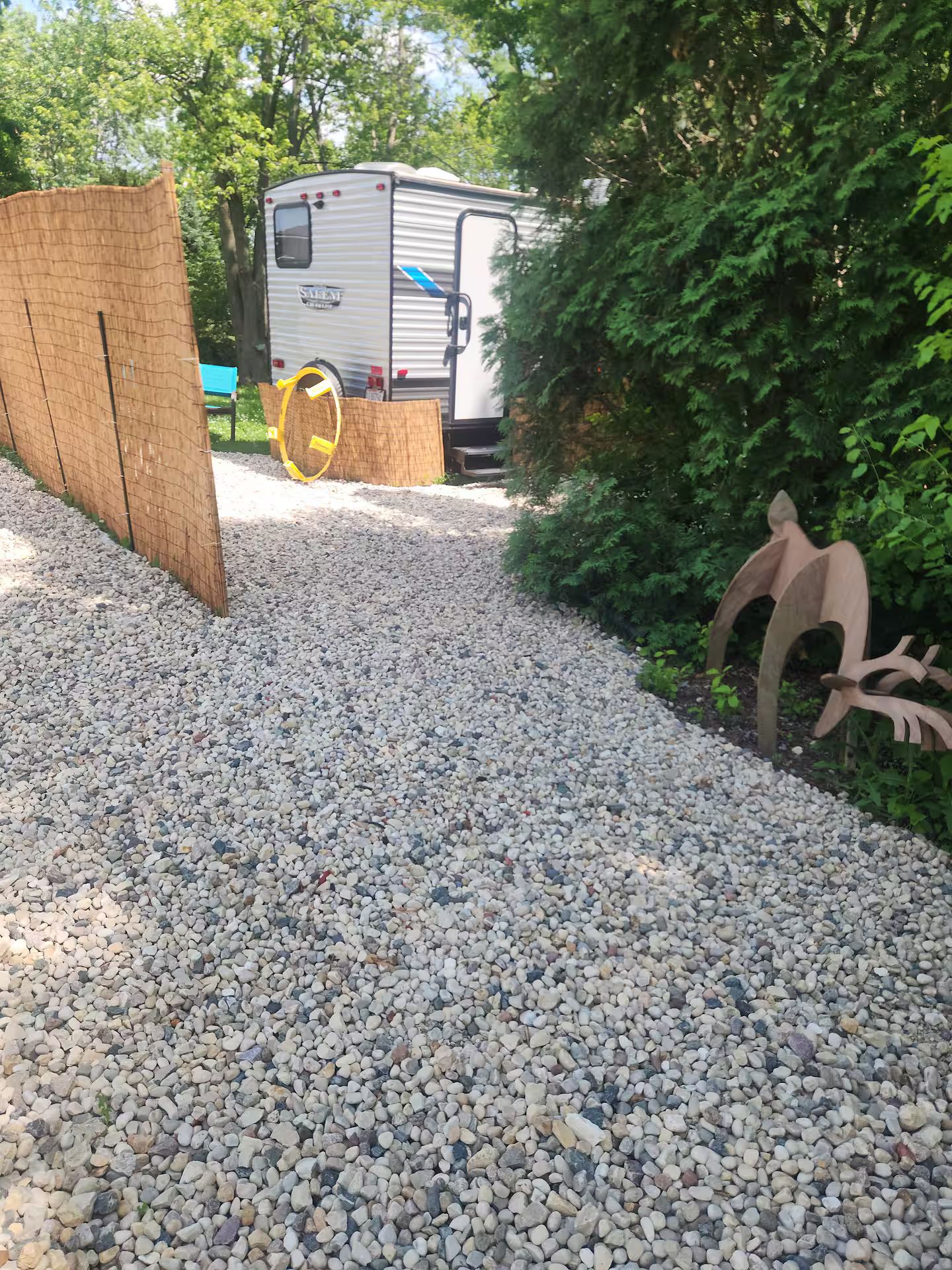 Ein Kiesweg führt zu einem Holztor, im Hintergrund steht ein weißer Wohnmobil und rechts davon eine Holzskulptur.