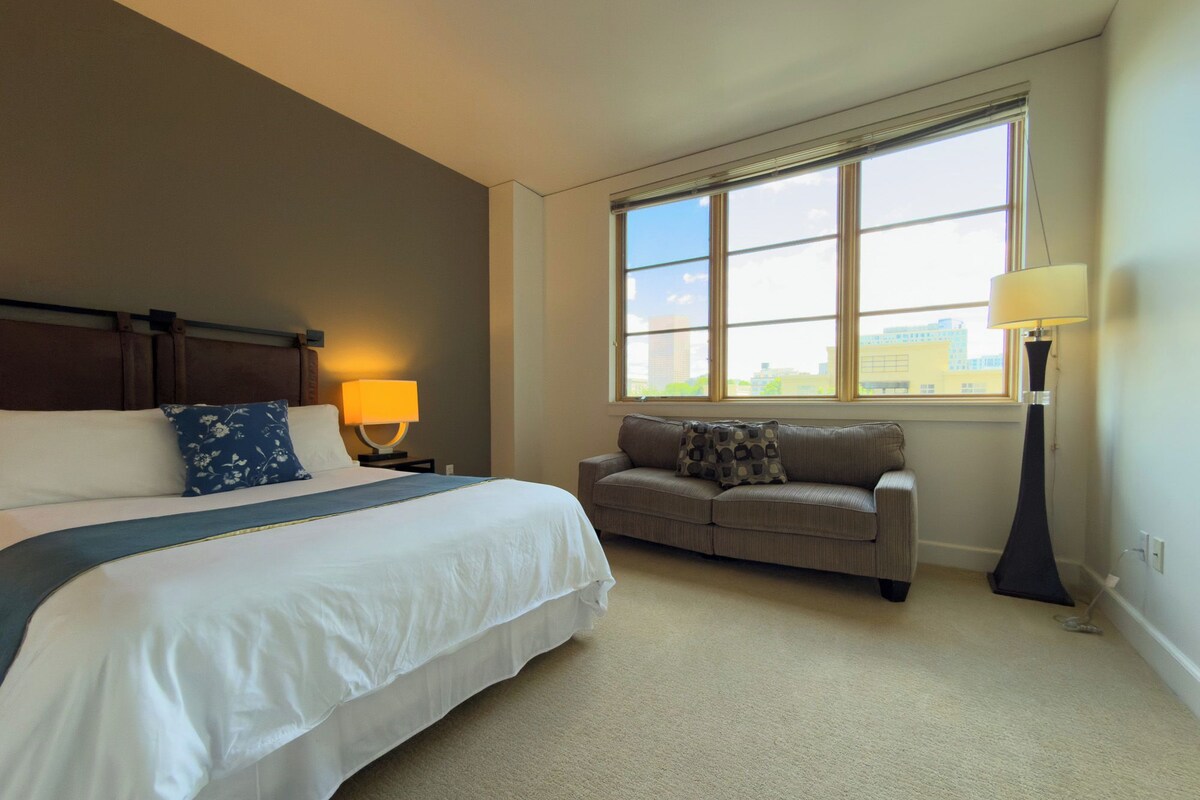 Una habitación de hotel con una cama grande, un sofá, un ventanal con vistas a la ciudad y una lámpara.