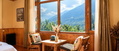 A room with wooden walls and ceiling, a large window with a mountain view, a bed, a table with chairs, and a radiator.