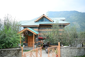 A two-story building with a stone facade and a metal roof, surrounded by greenery and a mountainous backdrop. - 3BHK Villa -That dreamy Stay in the Mountains of Naggar, Manali (Naggar)