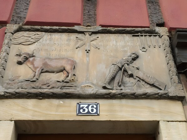 Un rilievo scolpito raffigura una mucca, una figura umana, un muro di pietra e un cartello con il numero 36.