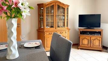 A dining area with a table set for two, a cabinet with glass doors, and a television on a stand.