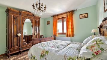 Une chambre avec une grande armoire en bois, un lit avec une literie à motifs floraux, un lustre et une fenêtre avec des rideaux.