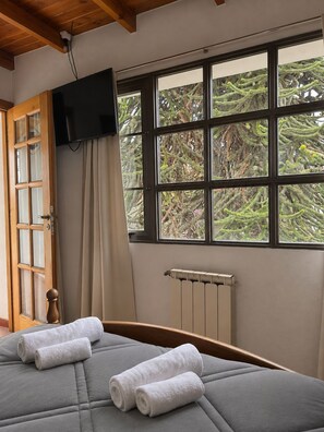 Una stanza con un letto, una televisione a muro, una finestra vista alberi e un termosifone.