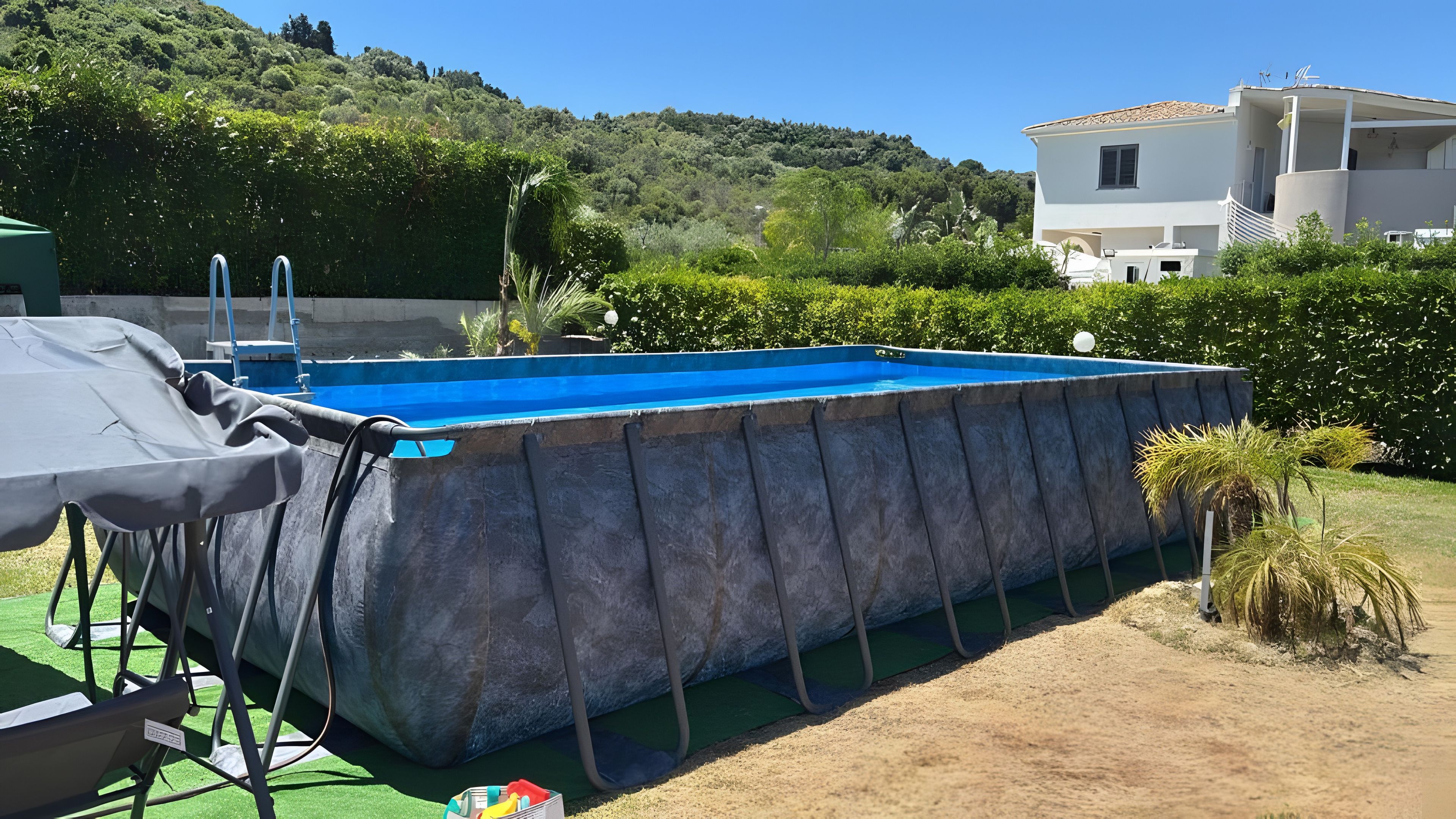 A swimming pool with a covered deck, surrounded by greenery and a hillside.