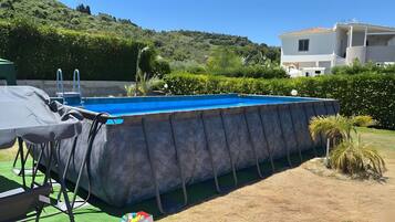 A swimming pool with a covered deck, surrounded by greenery and a hillside.