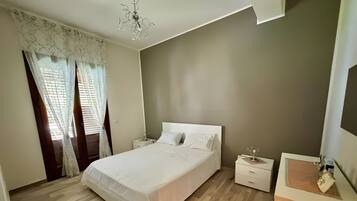A bedroom with a bed, bedside tables, a nightstand, a chandelier, and a window with curtains.