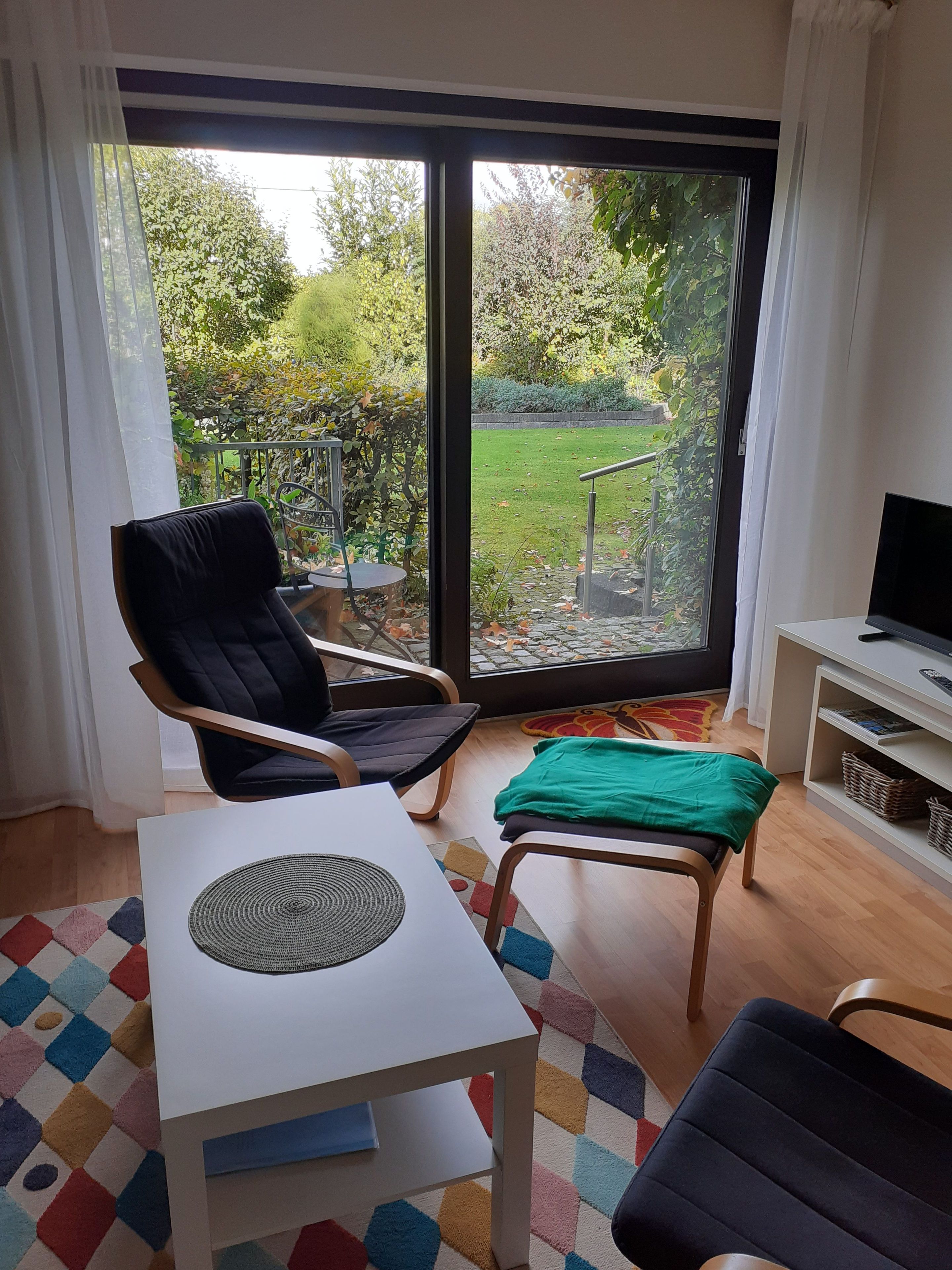Een kamer met een grote schuifdeur die uitkomt op een tuin, een zwarte stoel, een witte tafel en een televisie op een standaard.