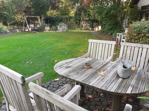 Een houten tafel en stoelen in een tuin met een gazon en een met bakstenen bestraat pad.