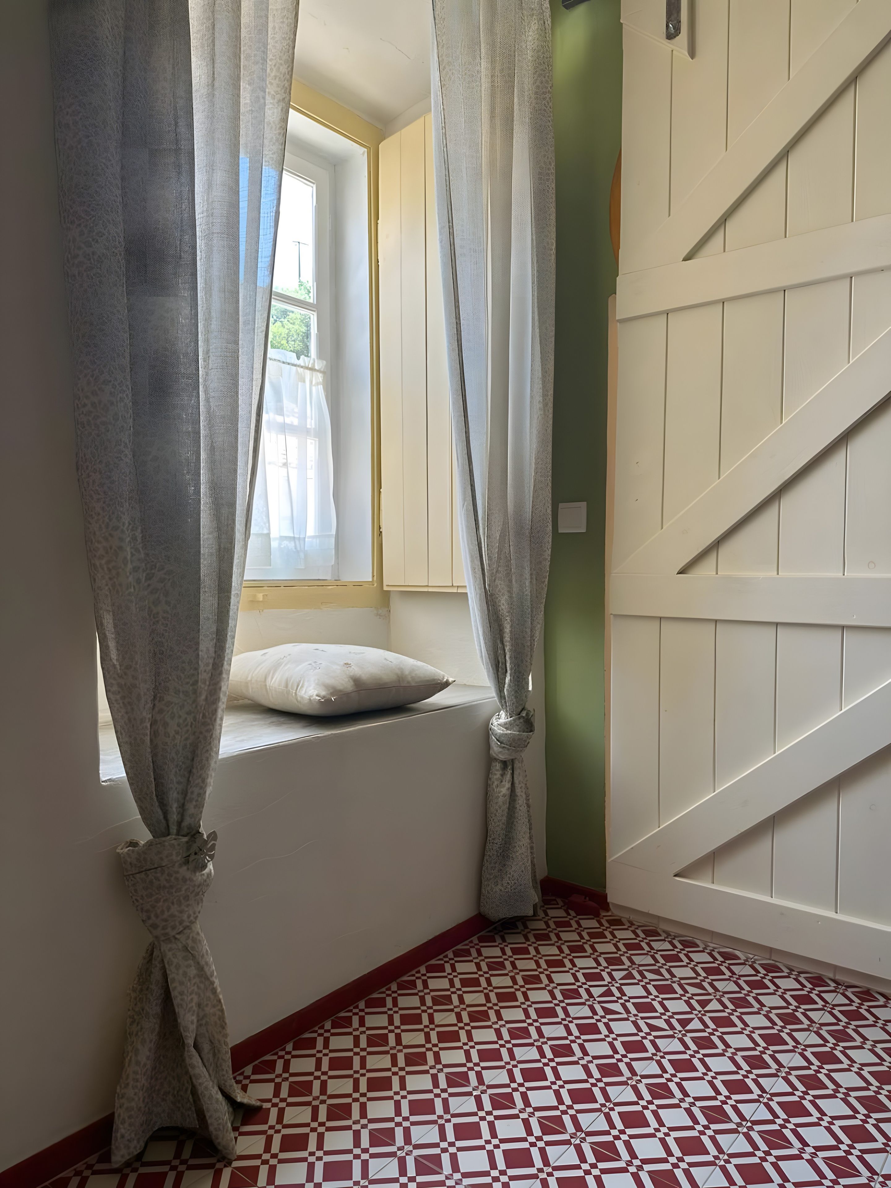 Una habitación con ventana, un almohadón en el alféizar, suelo estampado y una puerta blanca.