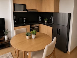Una cocina compacta con un refrigerador negro, un microondas y una cafetera. Hay una mesa redonda de madera con dos plantas en macetas.
