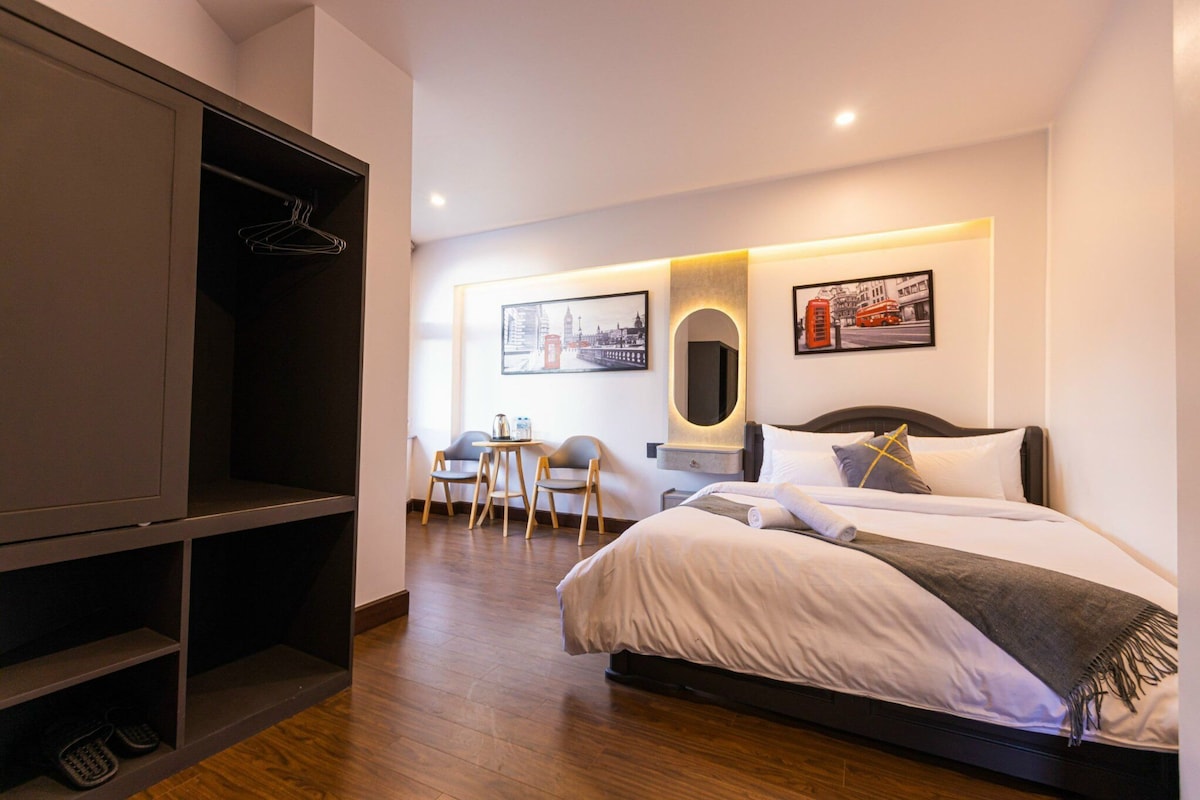 A hotel room with a bed, a wardrobe, a desk with chairs, and two framed pictures on the wall.