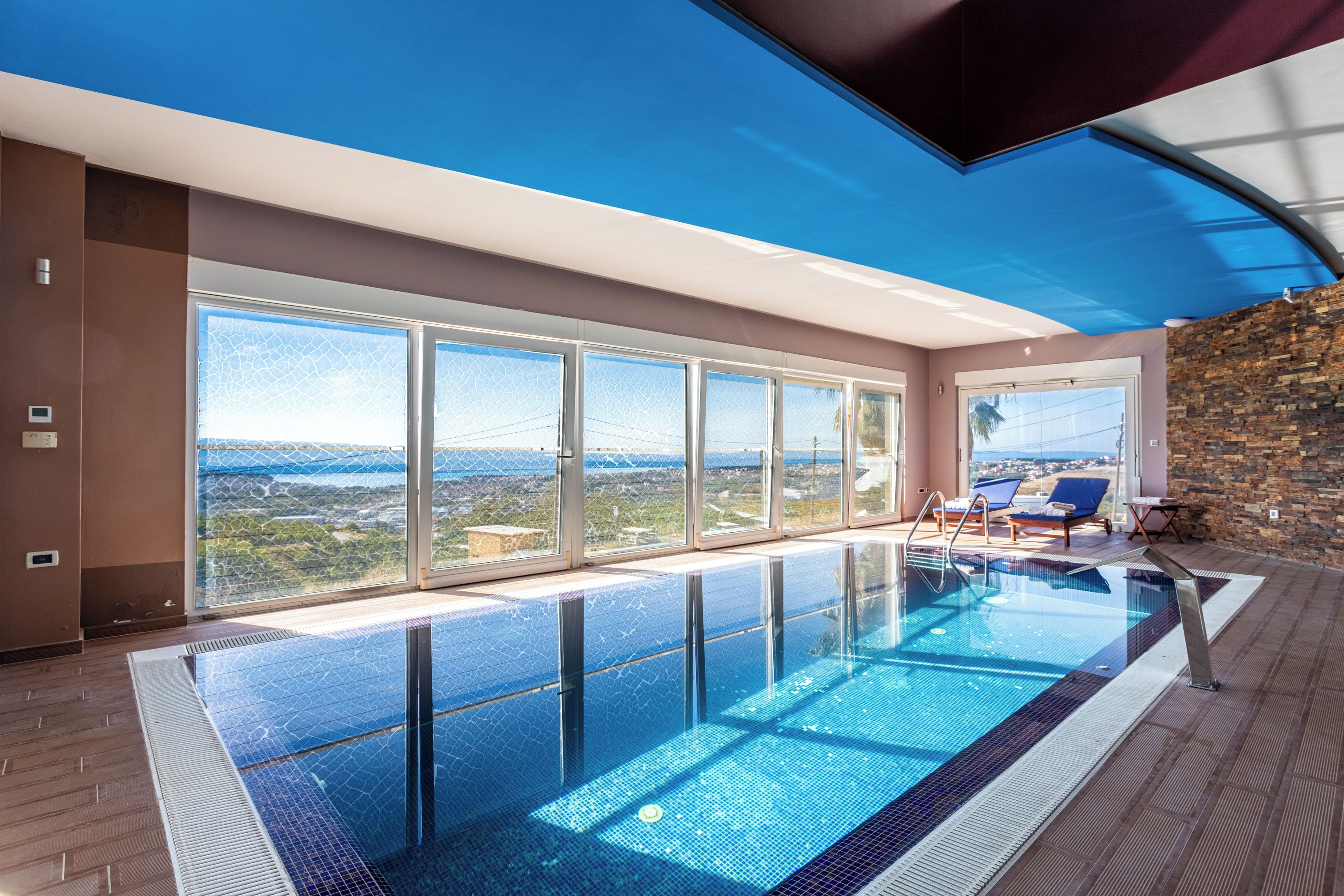 A modern indoor swimming pool with clear glass walls, a view of the landscape, and a stone wall.