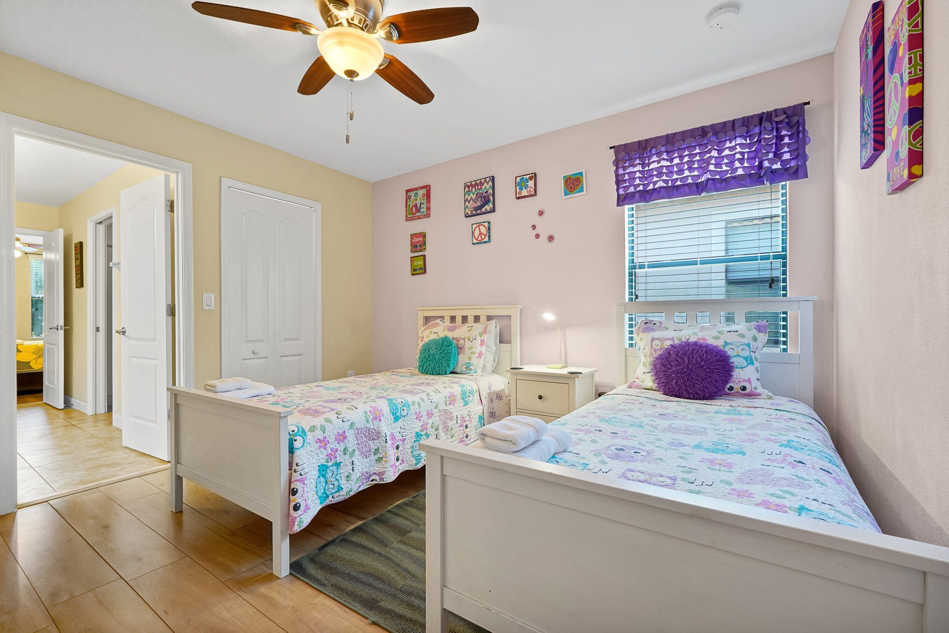 A bedroom with two single beds, a ceiling fan, a window with blinds, and a door leading to another room.