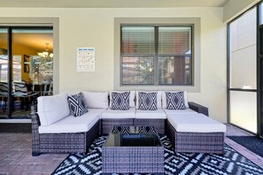A covered outdoor seating area with a white sofa and patterned cushions, a glass door entrance, and a window with blinds. - Just a Dream Away: 9-Bedroom Pool and Spa Home with Game Room (ChampionsGate)