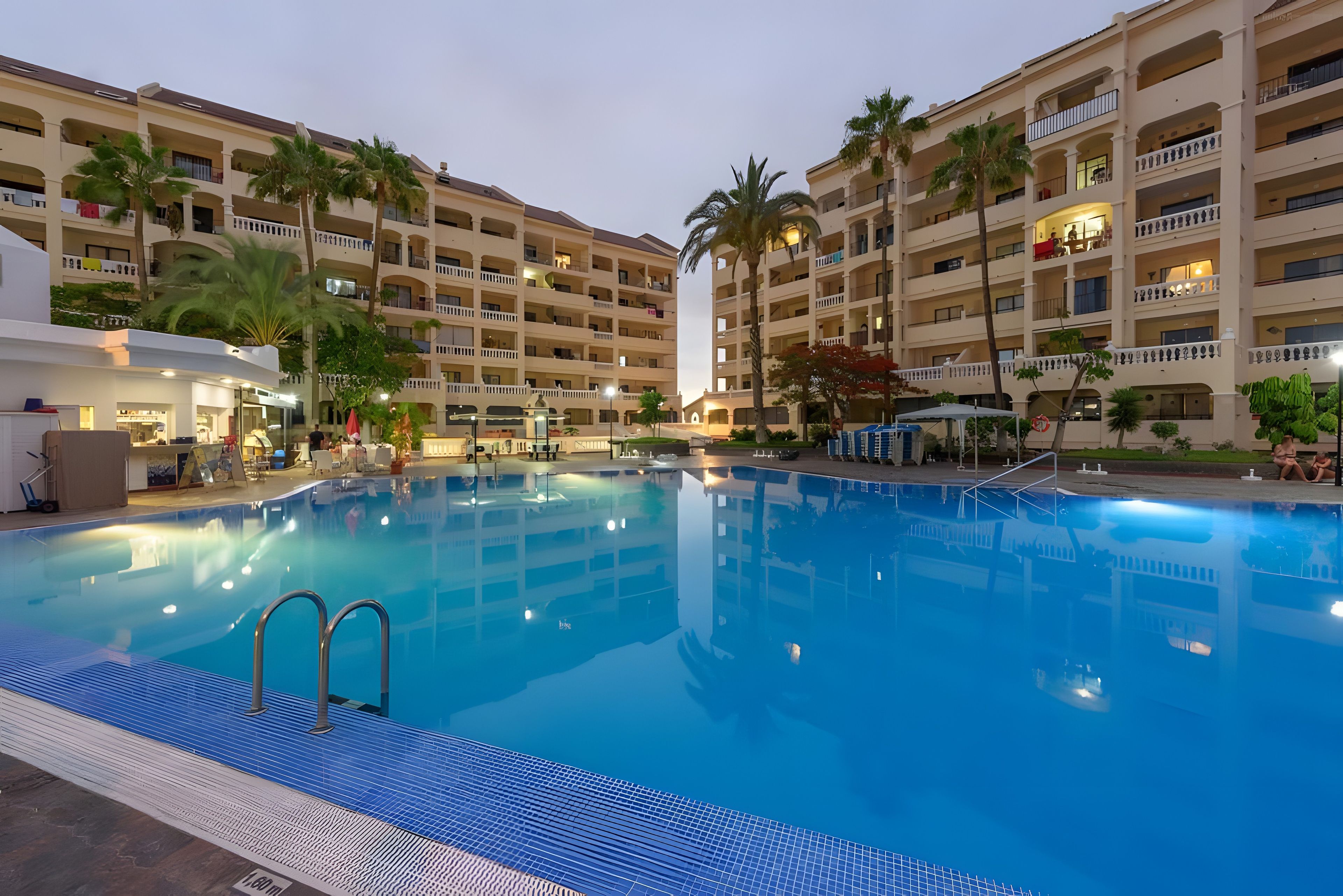 Een zwembadgedeelte van een hotel met meerdere balkons, palmbomen en een helderblauw zwembad.