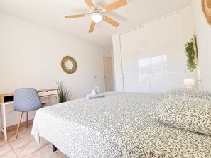 Een slaapkamer met een bed, een bureau, een stoel, een plafondventilator, een deur en een spiegel aan de muur.