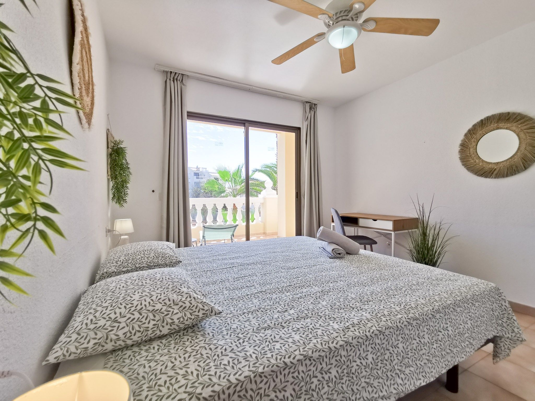 Une chambre avec un grand lit, un ventilateur de plafond, un bureau et une vue sur un balcon agrémenté de plantes.