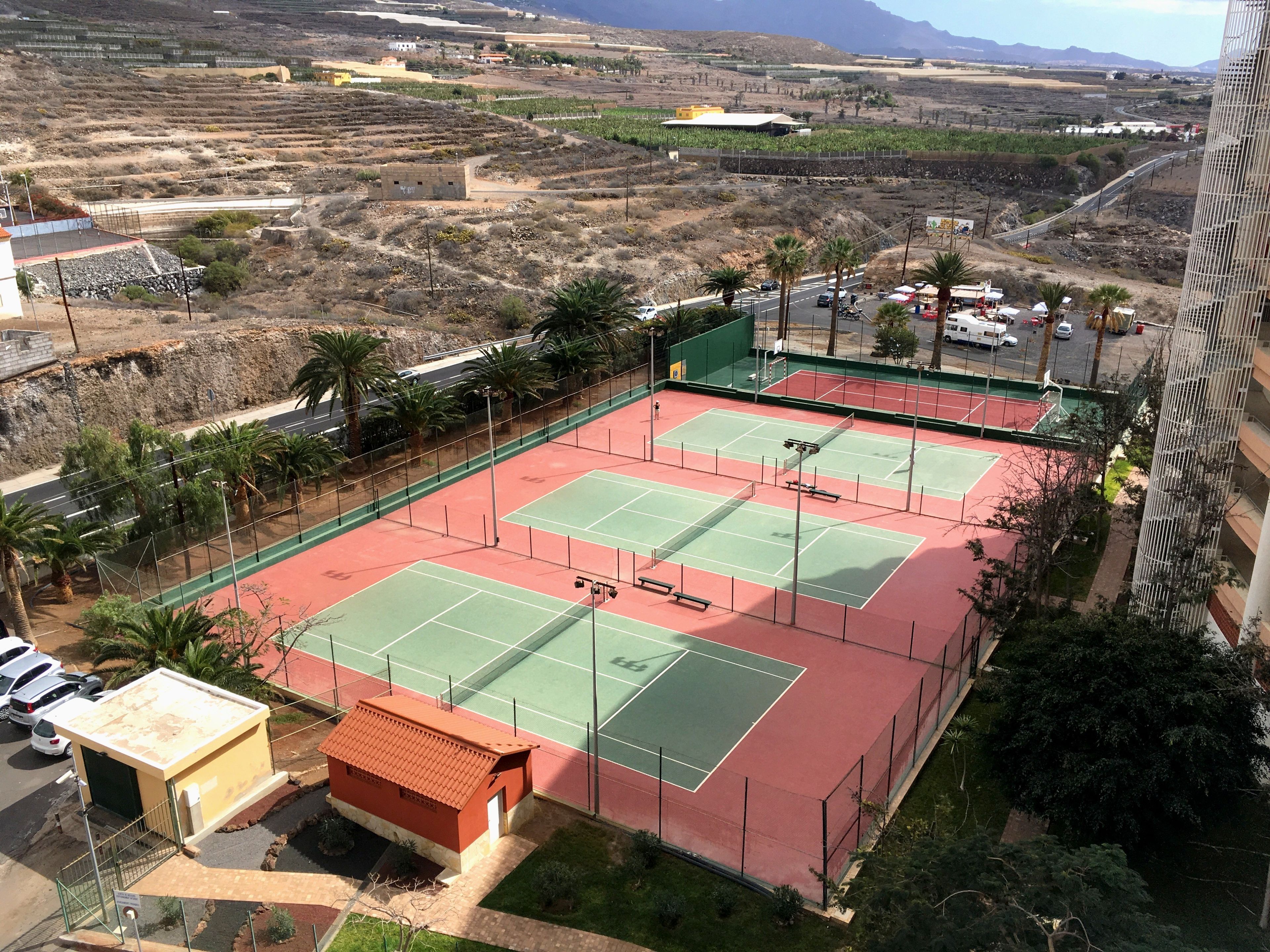 Een tennisbaan met een klein gebouw, palmbomen en een weg op de achtergrond.