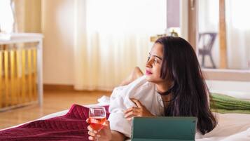 Una persona recostada en una cama con una computadora portátil y un vaso de líquido rojo, en una habitación con piso de madera y una ventana con cortinas.