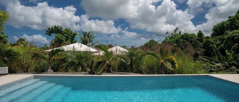 A clear blue swimming pool surrounded by lush greenery and a white building with a sloped roof.