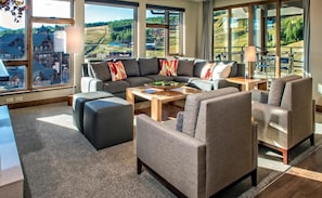 Un salon moderne comprenant un canapé d’angle, des fauteuils, une table basse et de grandes fenêtres donnant sur la campagne.