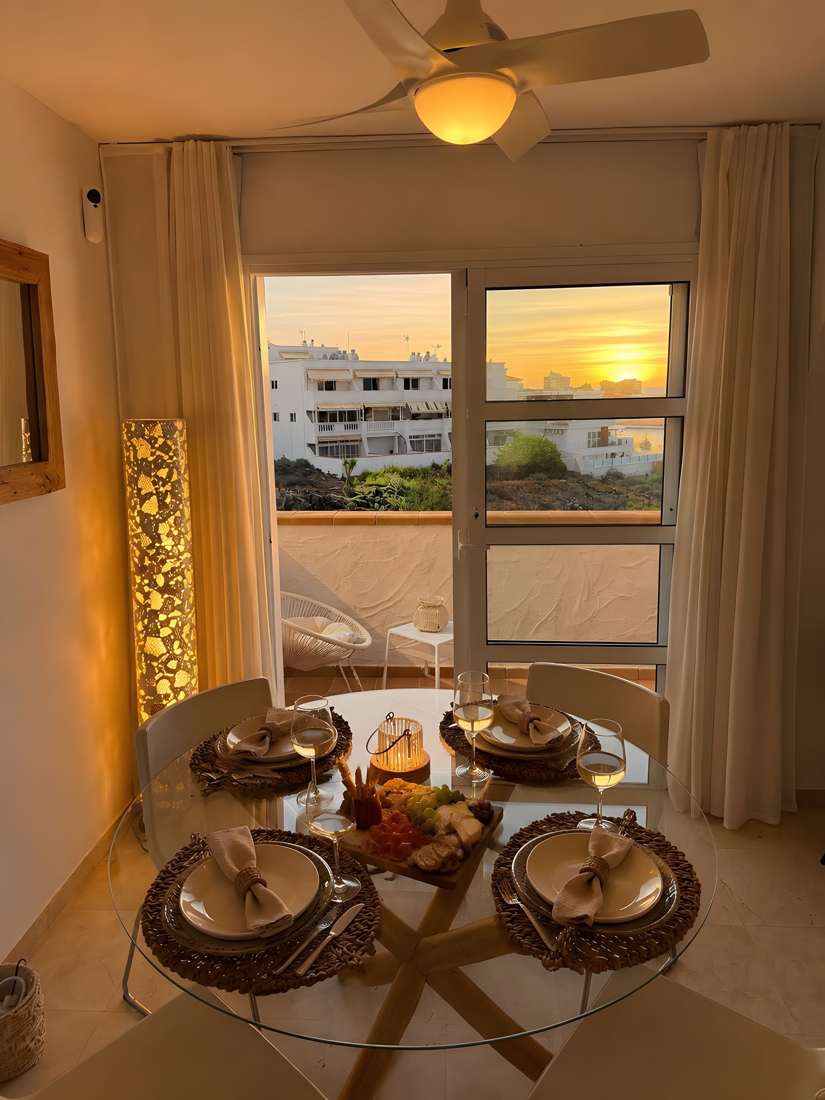 Un comedor con una mesa de vidrio dispuesta para cuatro personas, un ventilador de techo y vistas de edificios y el atardecer a través de la ventana.