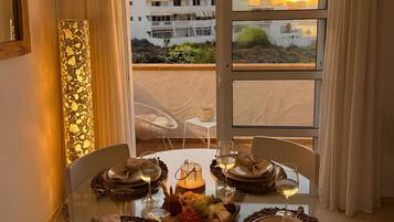 Un comedor con una mesa de vidrio dispuesta para cuatro personas, un ventilador de techo y vistas de edificios y el atardecer a través de la ventana.