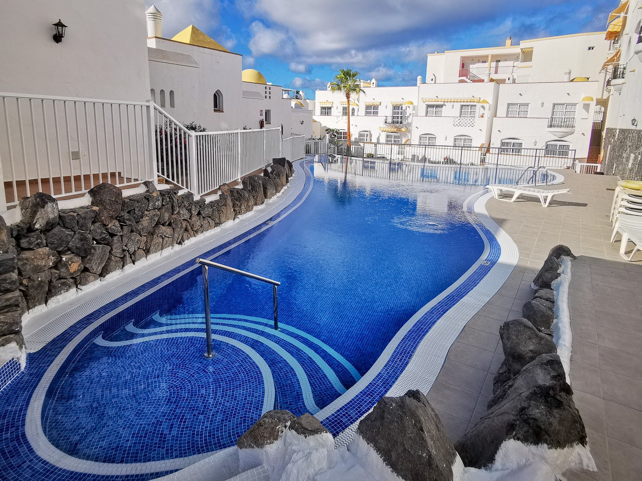 Una piscina con borde de azulejos, rodeada por un muro de piedra y edificios blancos con balcones.
