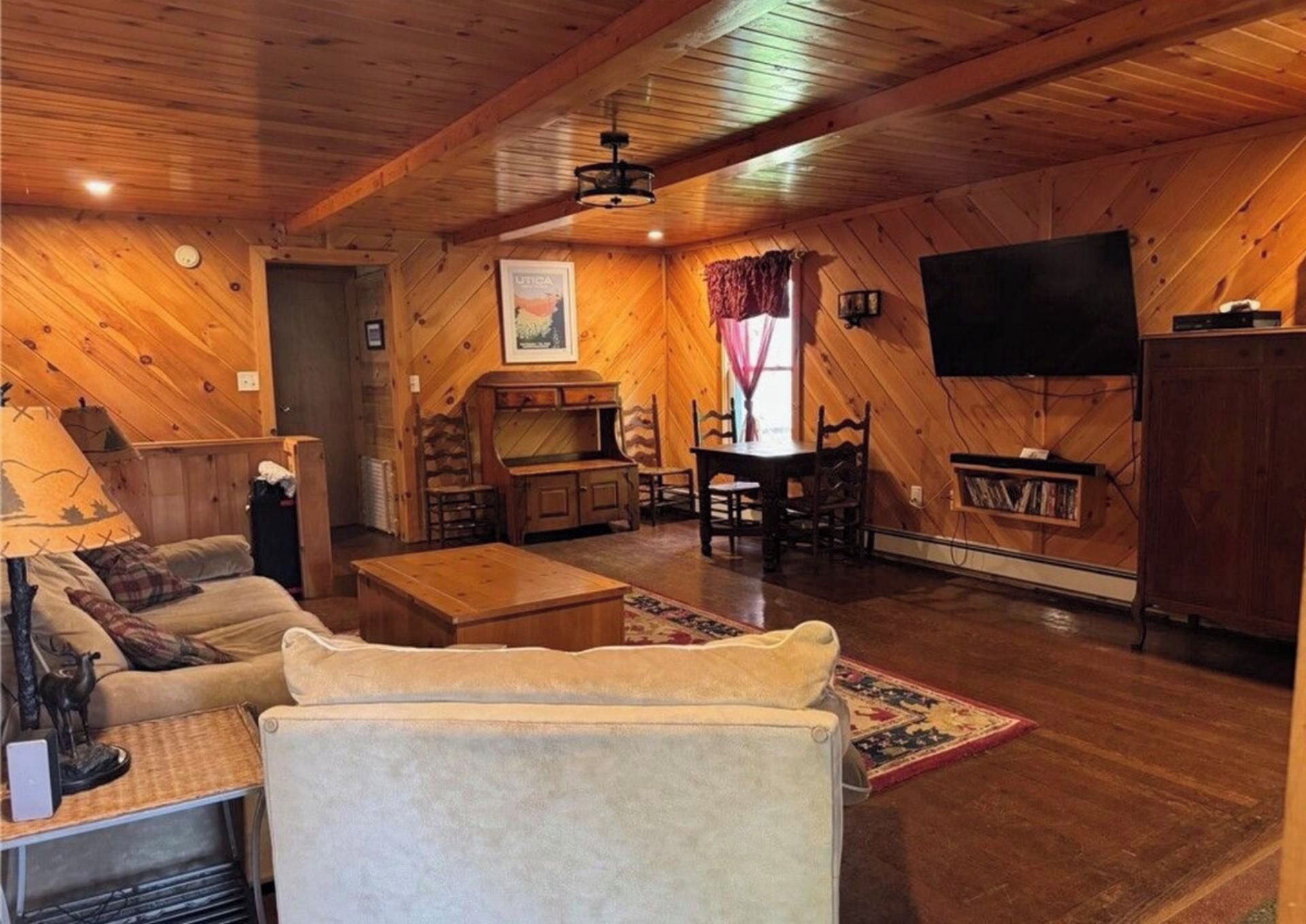 A cozy cabin interior with a wooden ceiling, a television mounted on the wall, a dining table with chairs, and a sofa.
