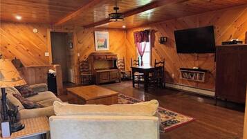 A cozy cabin interior with a wooden ceiling, a television mounted on the wall, a dining table with chairs, and a sofa.
