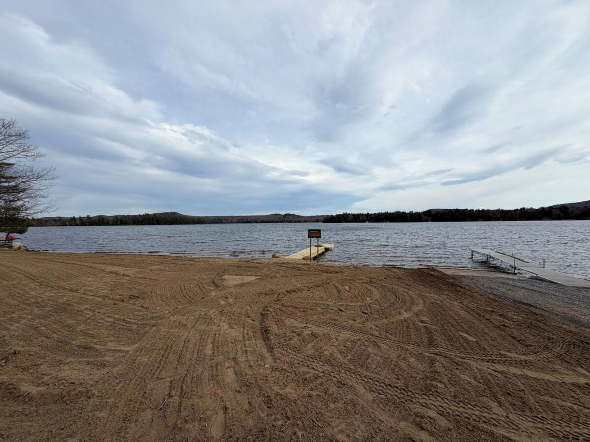A sandy beach area with a wooden dock extending into a lake, surrounded by trees and hills.