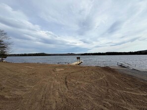 Una zona de playa arenosa con un muelle de madera que se adentra en un lago, rodeado de árboles y colinas.