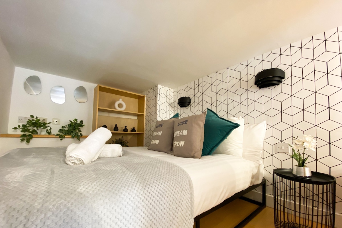 Una cama bien tendida con una manta blanca texturizada, una mesita de noche de madera con una lámpara negra y un papel tapiz con estampado geométrico.