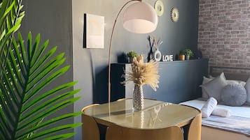 Una sala de estar moderna con una mesa redonda de vidrio, una lámpara de pie, una cama con un muro de ladrillo de fondo y una planta en maceta.