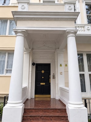 Una entrada de edificio con puerta negra, columnas blancas y una escalera de mármol.