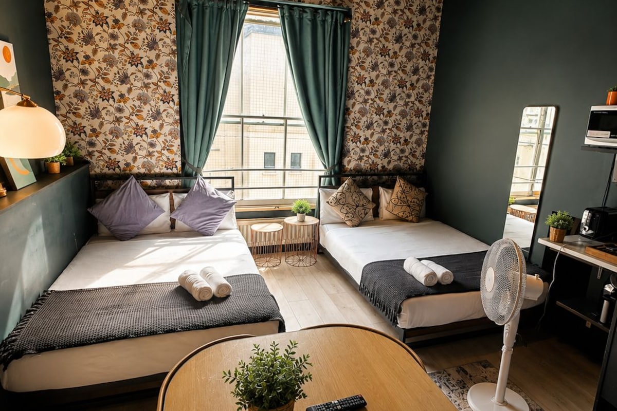 Una habitación acogedora con dos camas, una mesita con una planta, un ventilador de pie y un espejo en la pared.