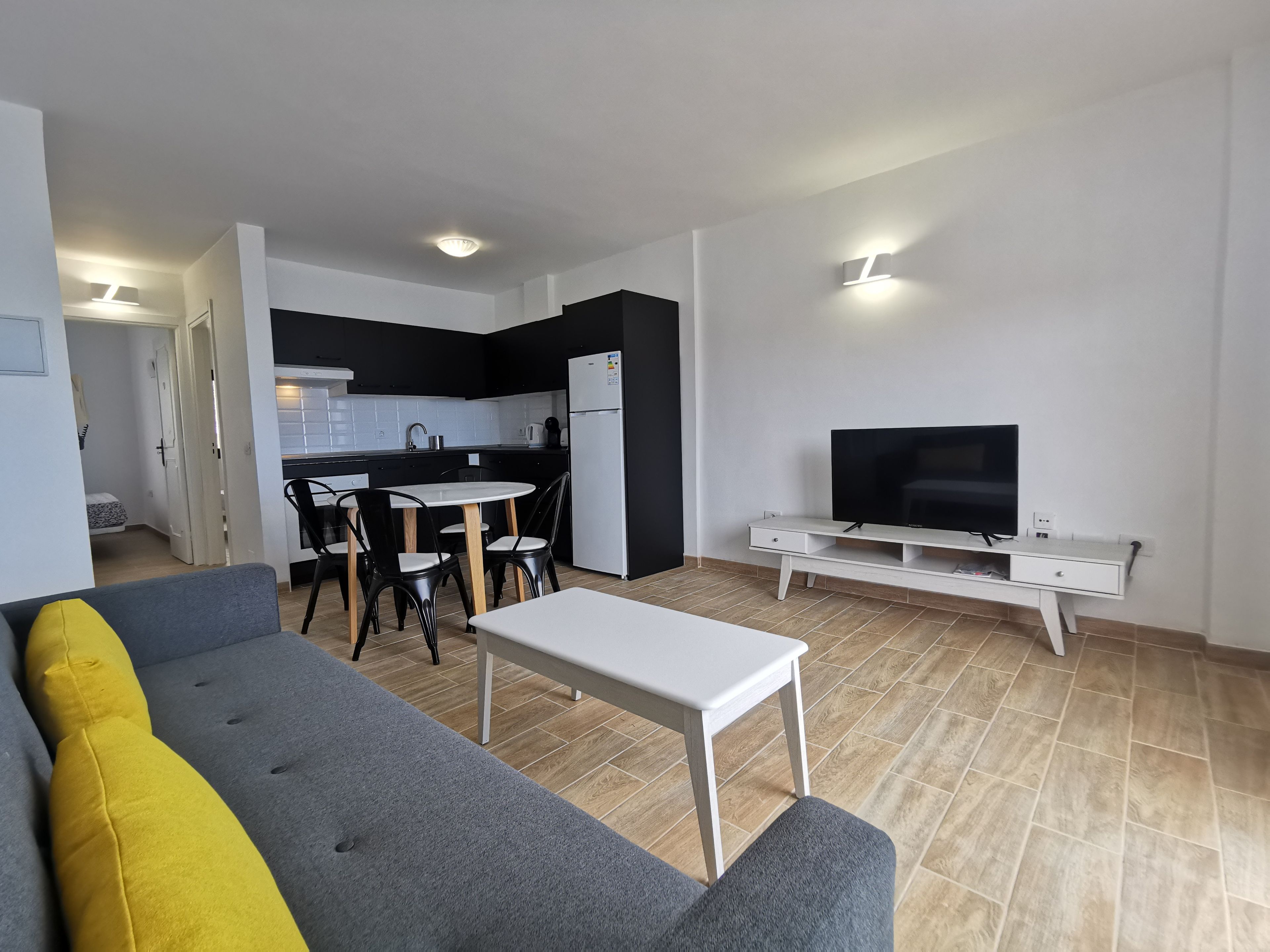 Een moderne woonkamer met een grijze bank, een klein wit tafeltje en een televisie op een witte dressoir.