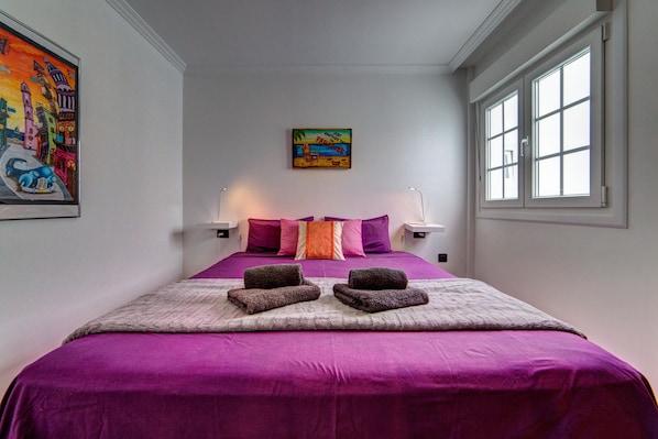 Een slaapkamer met een groot bed, twee bedlampen, een schilderij aan de muur en een raam met zonwering.
