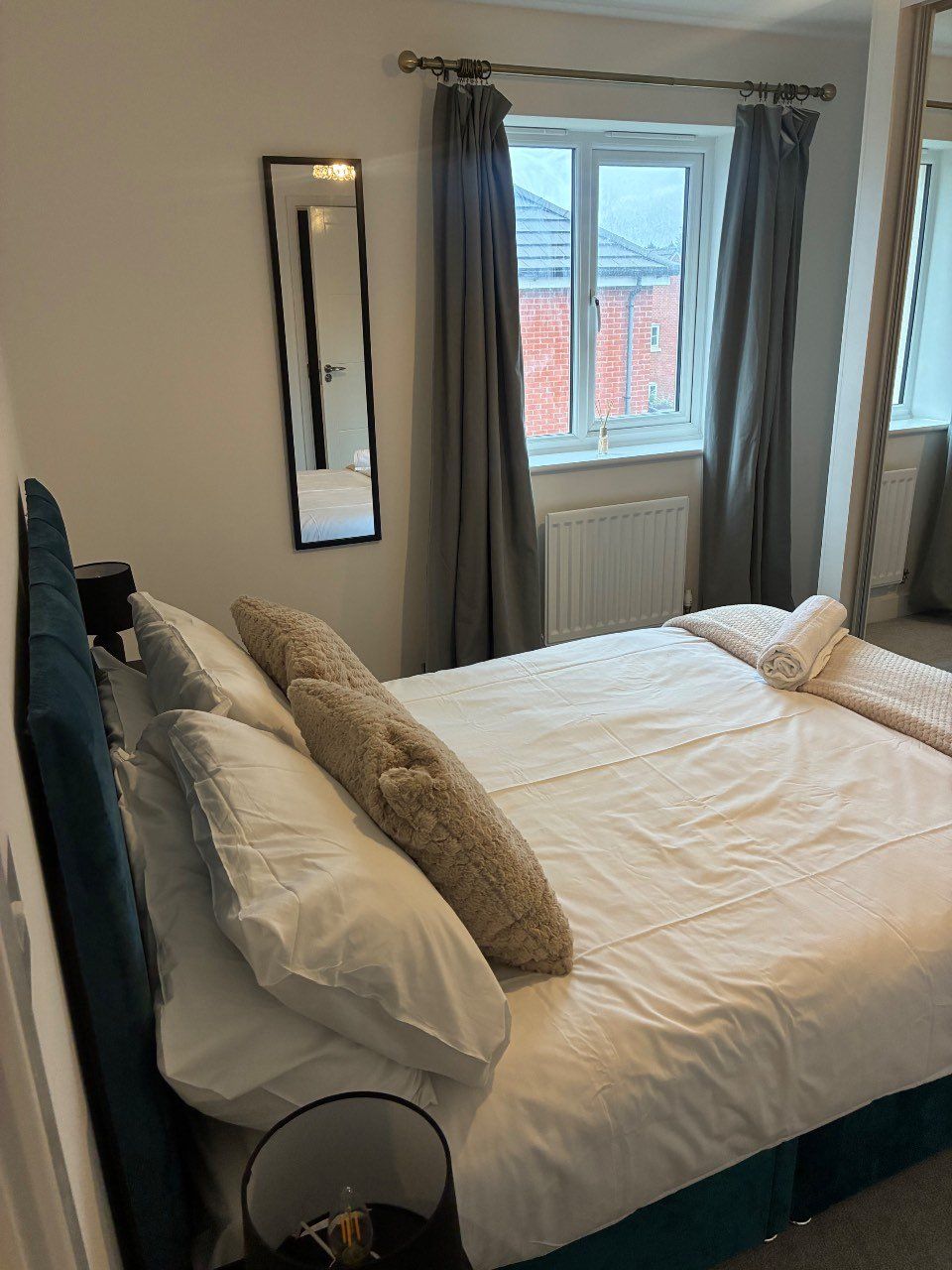 A bedroom with a bed, pillows, a bedside table, a mirror, and a window with curtains.