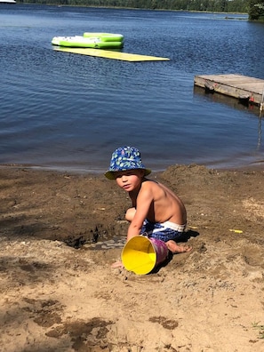 Un enfant joue avec un seau jaune sur une plage de sable, près d’un lac paisible. On aperçoit en arrière-plan un quai et des bateaux gonflables.