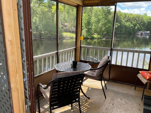 Un espace extérieur abrité, doté d’une table et de chaises, qui offre une vue sur un lac avec un quai et des bâtiments en arrière-plan.