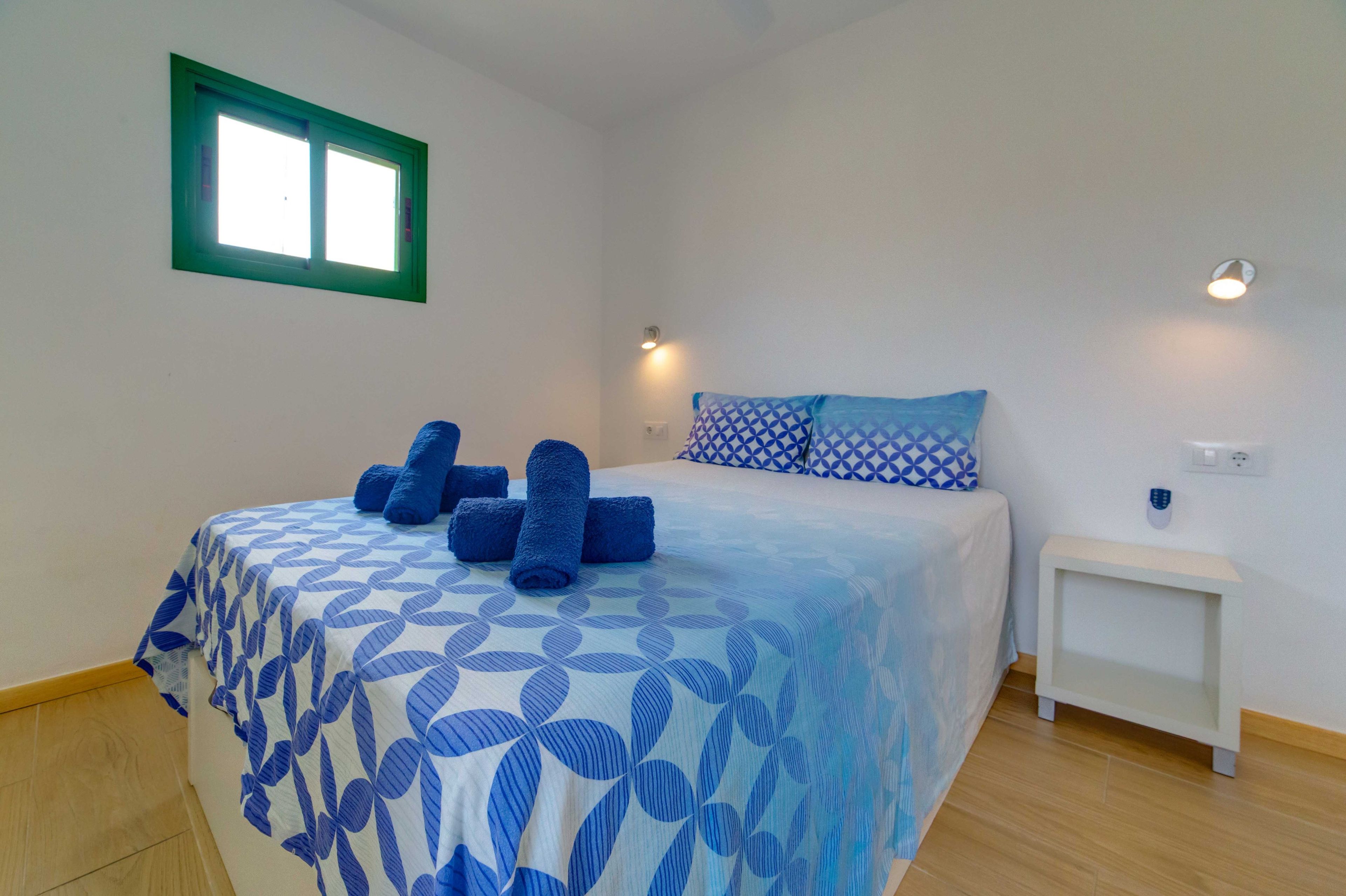 Een slaapkamer met een bed, kussens, een nachtkastje, een wandlamp en een raam met een groene omlijsting.