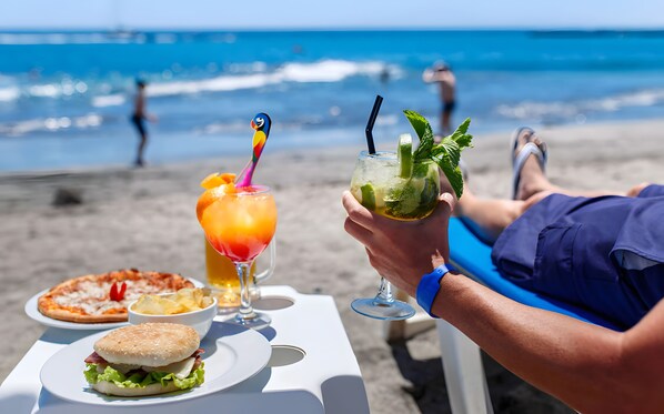 Une table installée sur la plage, avec de la nourriture et des boissons, d’où l’on a vue sur l’océan.