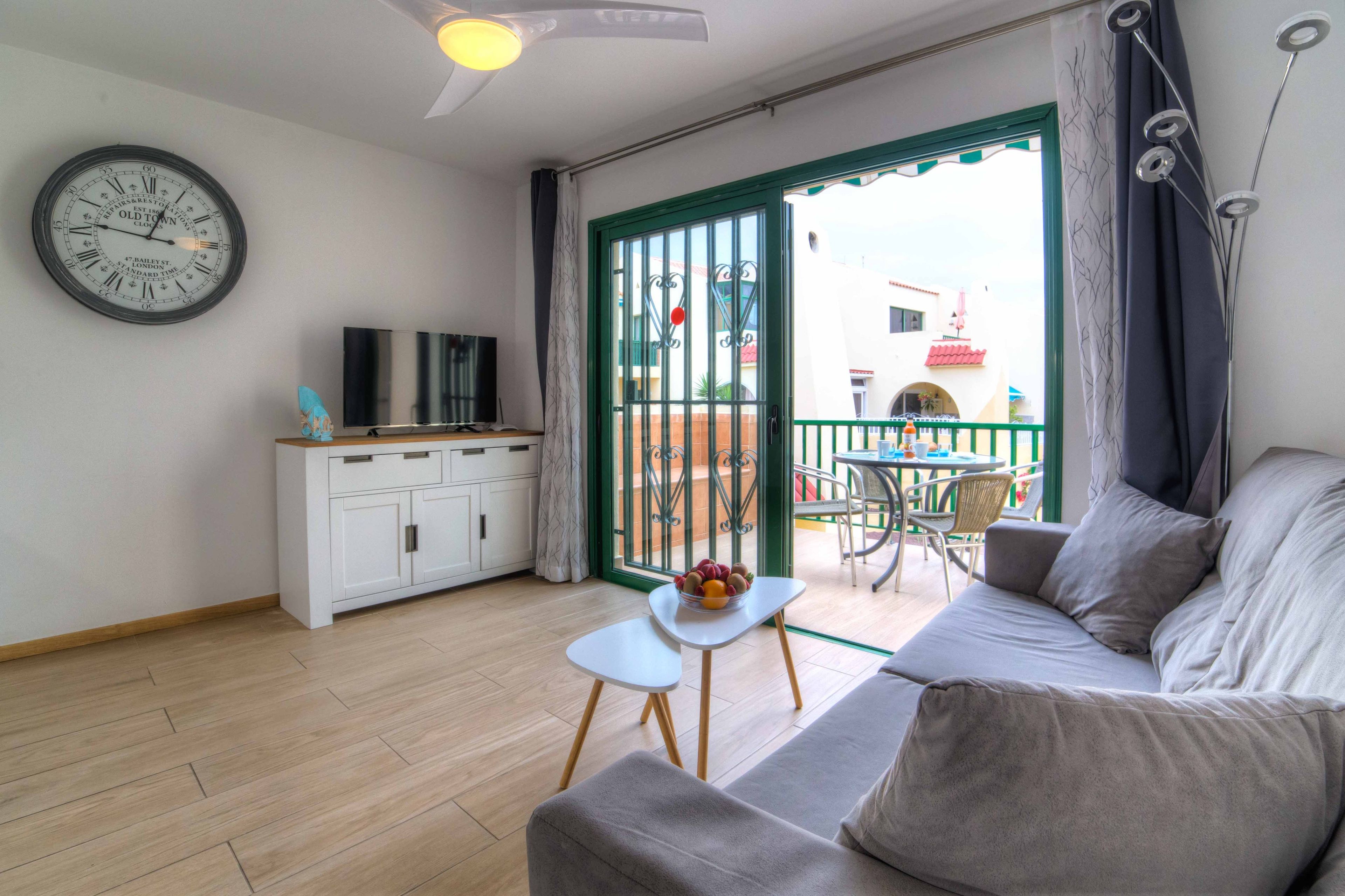 Een woonkamer met een bank, een televisie, een klein tafeltje met fruit en een balkon met een tafel en stoelen.