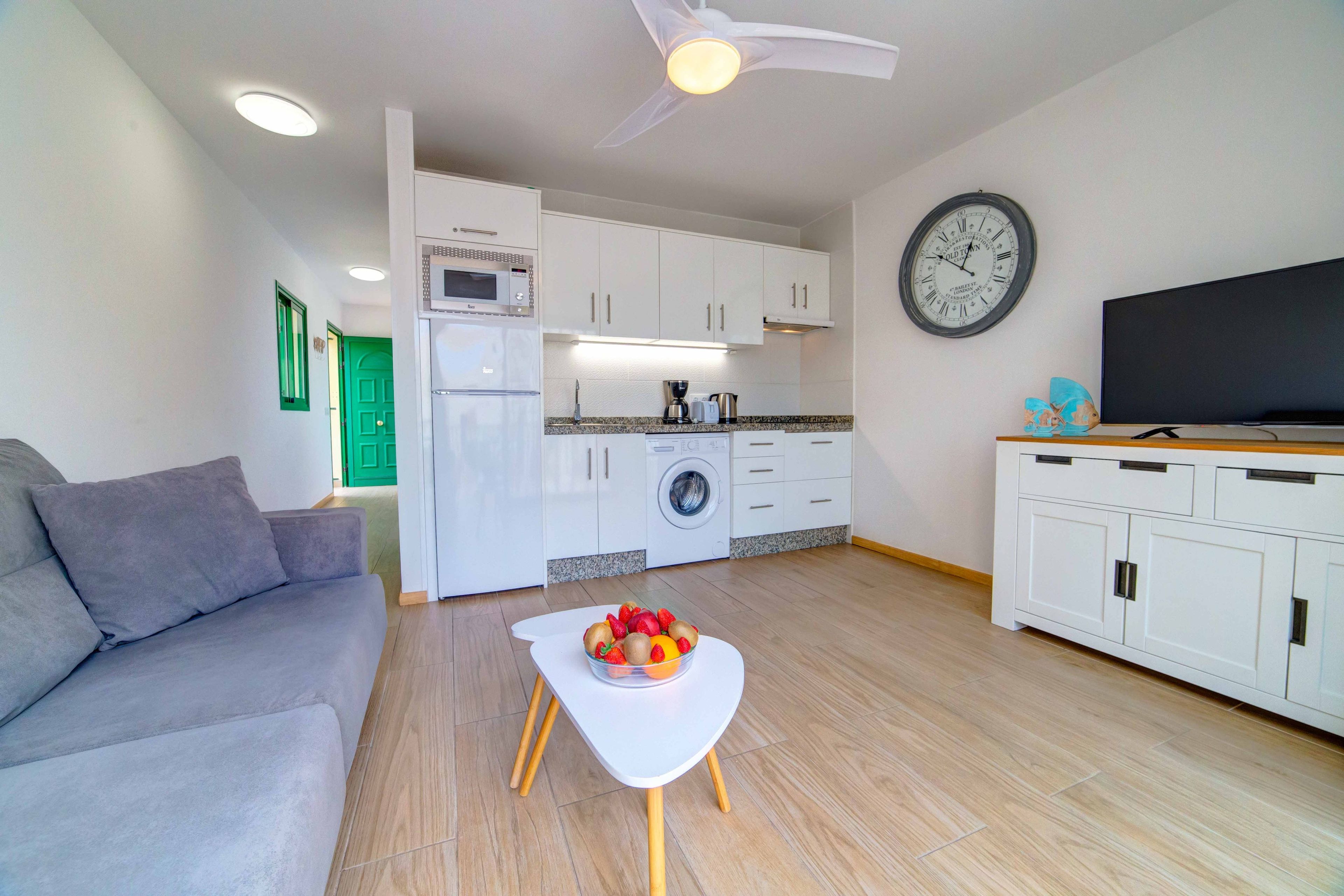 A compact living space with a kitchenette, a dining area with a fruit bowl, a sofa, and a television on a cabinet.