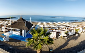 Een hotel aan het strand met meerdere ligbedden en parasols, een rieten constructie en een palmboom.