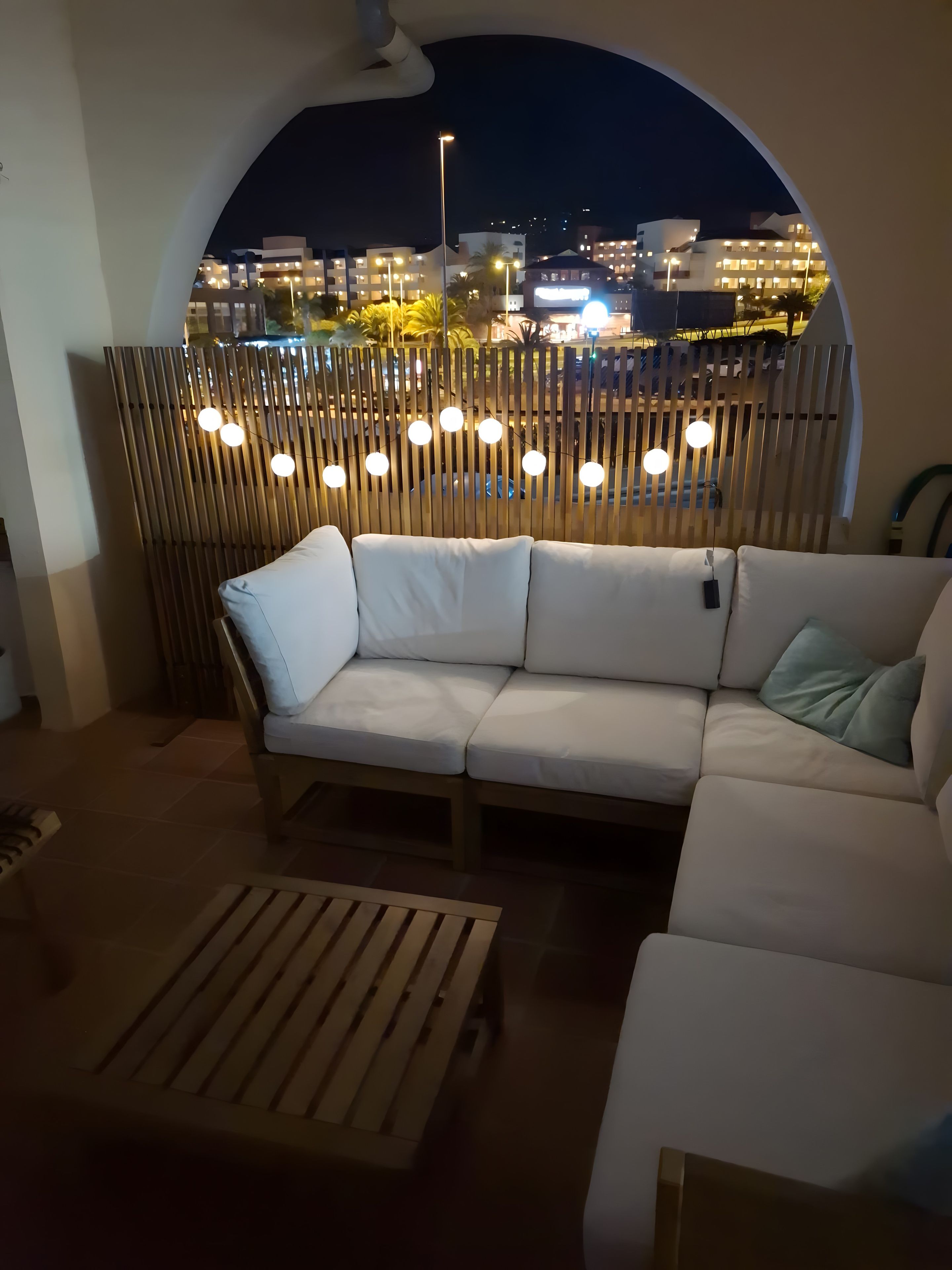 Et moderne udendørs opholdsområde med en hvid sofa og et træbord, med udsigt til en by om natten.