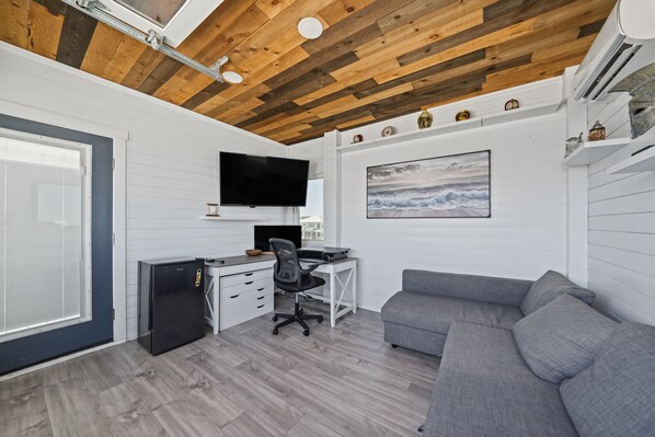 A compact living space with a wooden ceiling, a flat-screen TV mounted above a desk, a gray sofa, and a small refrigerator. - The Sweet Spot- Upstairs Apartment (Carolina Beach)